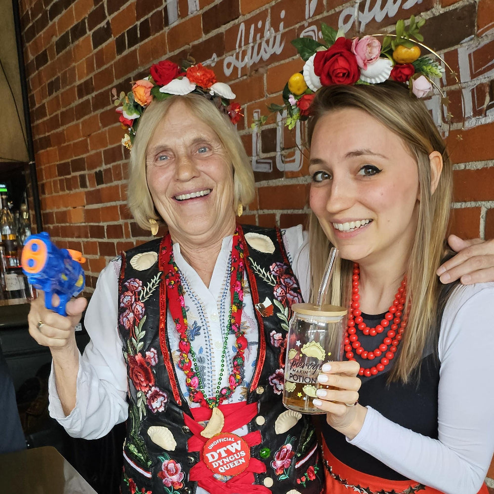 Two women in traditional Polish attire with floral and pierogi headpieces, one holding a drink and water gun, against a brick on Dyngus Day in Ohio.